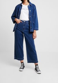 Žena v tmavě modrých volných džínech a odpovídající denimové bundě přes bílém tričku "Tommy Jeans" s černými vysokými teniskami.