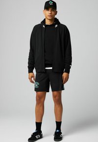Schwarze Zip-up Kapuzenjacke mit weißen Streifen, schwarzer Sweatshirt, schwarze Shorts mit grünem Text, schwarze Socken und schwarze Schuhe. Schwarze Kappe mit Logo.