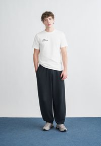 Balti trumpomis rankovėmis marškinėliai su juodu „Nike Official“ logotipu, derinami su laisvomis juodomis sportinėmis kelnėmis ir šviesiai pilkais sportiniais batais.