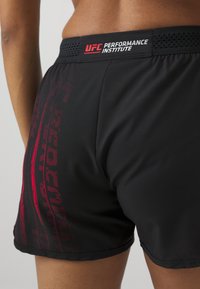 Gros plan sur une personne portant un short noir UFC Performance Institute avec des graphismes rouges sur le côté, montrant le bas du dos et le haut des jambes.