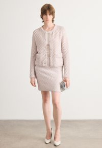 Lichtroze tweed jasje met een gestructureerd patroon en twee voork huizen, gedragen met een aansluitende kokerrok. Het model houdt een zilveren clutch vast.