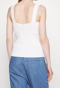 Canottiera bianca a costine con spalline larghe, caratterizzata da un design incrociato sul retro. Abbinata a pantaloni in denim a gamba larga di colore azzurro chiaro.