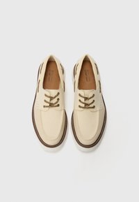 Beige canvas shoes with brown accents and laces, rounded toe, perforated design on the sides, and contrasting stitching along the sole.