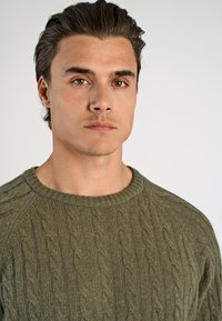 Groene kabelgebreide trui met een ronde hals. Heeft een textuurpatroon en ribbelmanchetten. Model toont het kledingstuk tegen een neutrale achtergrond.
