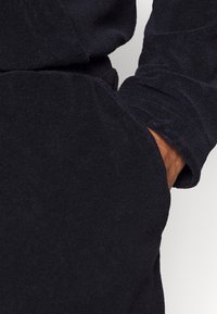 Tissu texturé bleu marine foncé montrant une main dans une poche. Le tissu semble doux avec une finition moelleuse, présentant un détail de manchette au niveau de la manche.