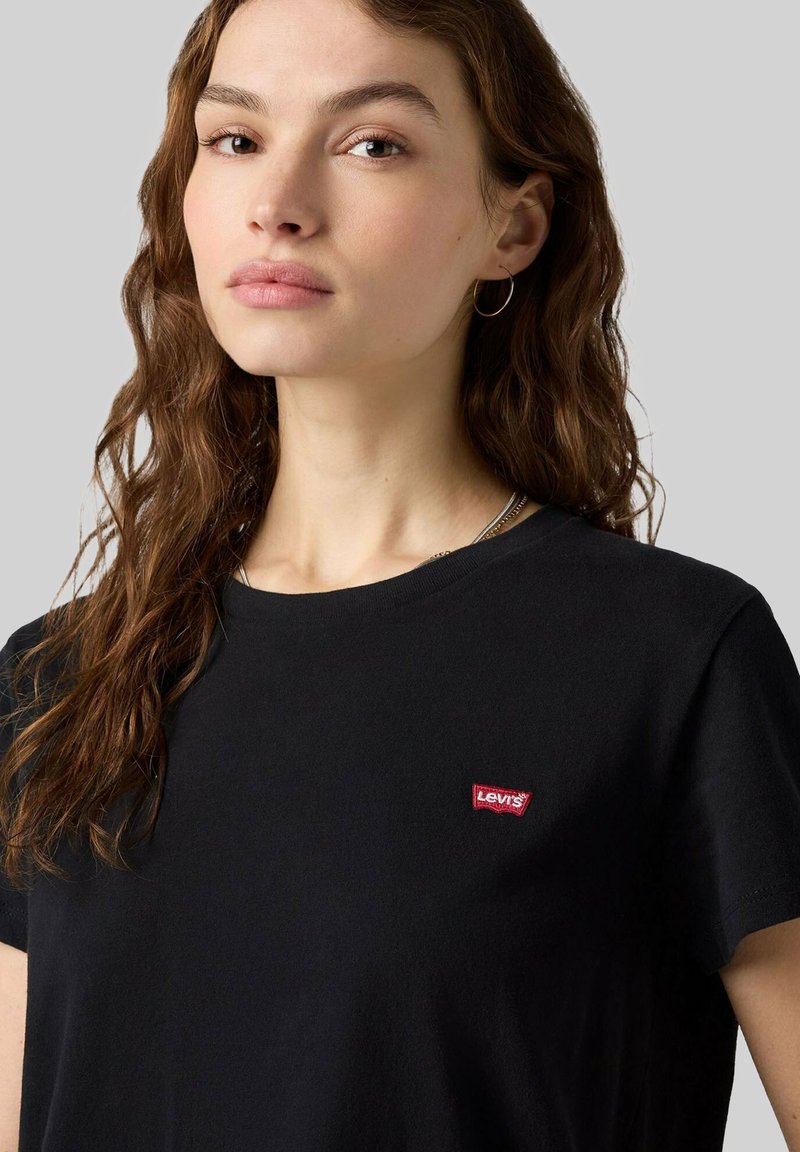 Junge Frau mit welligem braunem Haar trägt ein schwarzes Levi’s T-Shirt und einen silbernen Creolen-Ohrring, schaut leicht zur Seite vor einem schlichten grauen Hintergrund.