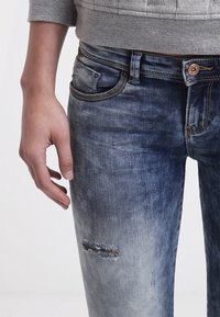 Jeans de mezclilla con un lavado azul desgastado, detalle de rodillas desgastadas, diseño de cinco bolsillos y costuras en contraste en la cintura.