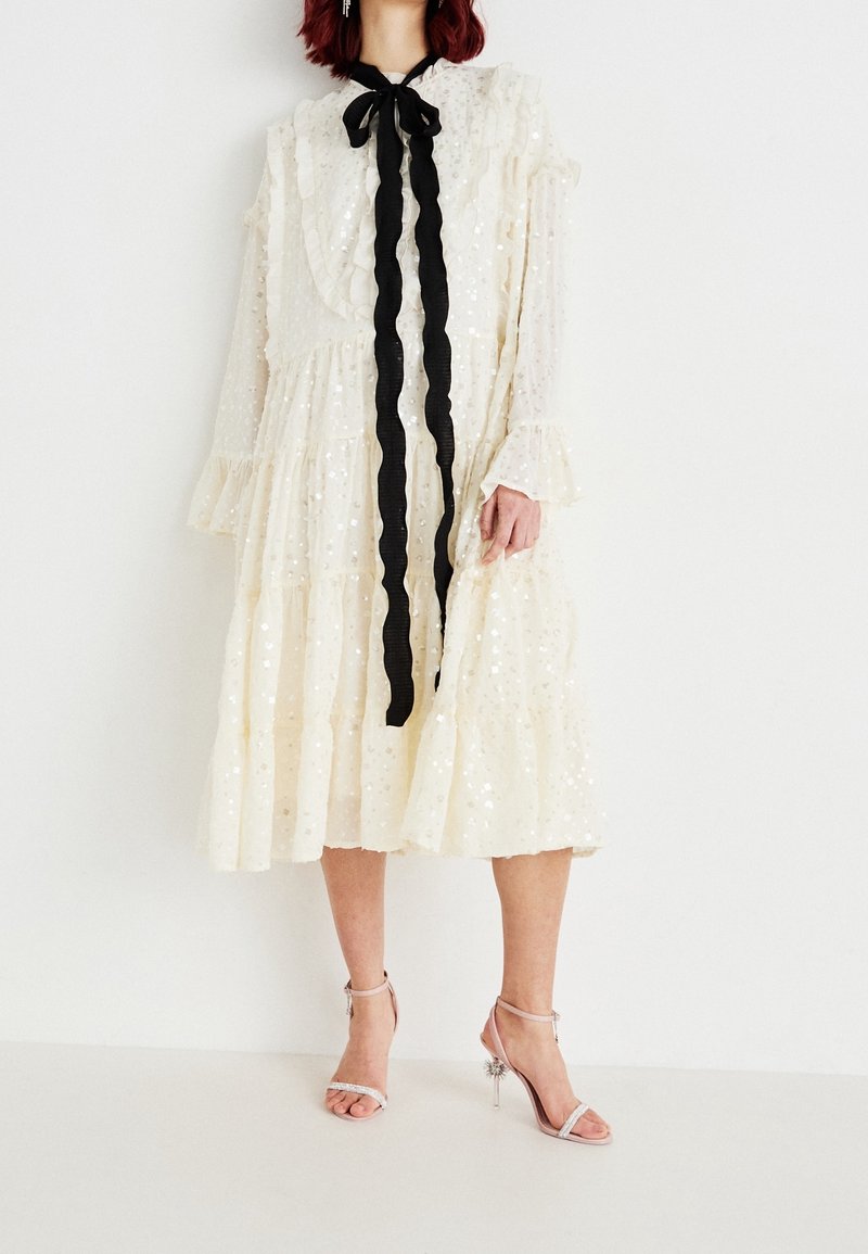 Femme portant une robe midi blanche à manches longues avec des sequins, des volants et un long nœud papillon noir, associée à des sandales argentées à talons hauts.