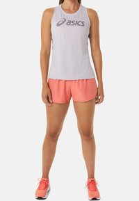 Mujer con una camiseta sin mangas blanca de Asics, pantalones cortos de running coral y zapatillas de running coral, de pie con los brazos relajados a los lados.