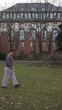 Άνδρας που φορά γυαλιά ηλίου περπατά στο γρασίδι μπροστά από ένα μεγάλο κτίριο από κόκκινα τούβλα με αψιδωτά παράθυρα και δέντρα χωρίς φύλλα.