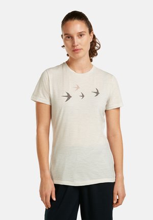 Kobieta z brązowymi włosami związanymi do tyłu, ubrana w jasnobeżowy t-shirt z czterema sylwetkami lecących ptaków na wysokości klatki piersiowej.
