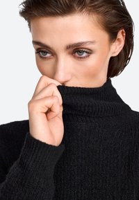 Schwarzer Rollkragenpullover aus strukturiertem Strickstoff. Der hohe Kragen und die Handgelenkabschlüsse haben gerippte Akzente. Die Hand des Models ist nah am Kragen.