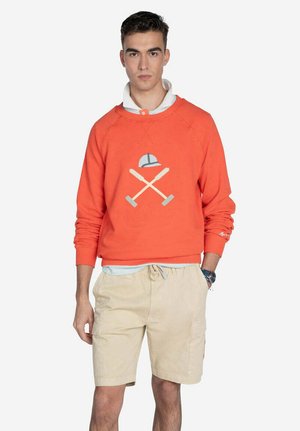 Sudadera naranja con cuello redondo, que presenta un diseño gráfico de un sombrero y remos cruzados. Combinada con pantalones cortos beige.