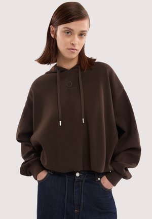 Junge Frau mit glattem braunem Haar, die einen dunkelbraunen Trussardi-Hoodie und dunkelblaue Jeans trägt, Hände in den Taschen, neutraler Hintergrund.