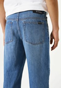 Jean en denim bleu clair avec une coupe décontractée, doté de poches arrière et d'une étiquette de marque noire sur la ceinture. Détail de couture distinctif.
