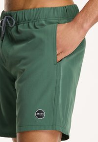 Zelene kupaće gaće izrađene od laganog materijala, s elastičnom waistband-om, bočnom džepom i okruglom logom na rubu.