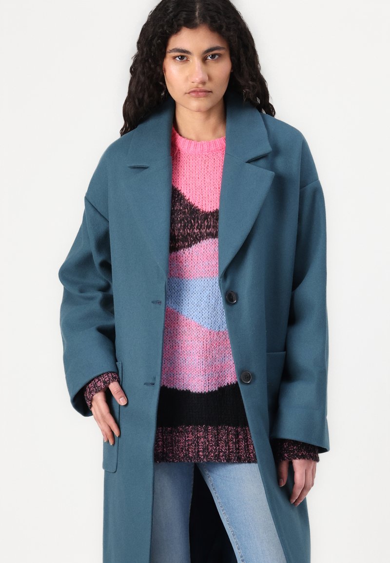 Türkiser Oversized-Mantel mit breiten Revers, doppelten Knöpfen und Seitentaschen, getragen über einem mehrfarbigen Strickpullover mit strukturierten Mustern.
