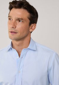 Lichtblauwe overhemdbloes met dunne verticale strepen, kraag en een enkele rode accent op de knooplijst. Glad, professioneel stoftextuur.