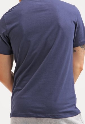 Mężczyzna w jednolitym granatowym t-shircie z krótkim rękawem i jasnoszarych spodniach, pokazany od tyłu z częściowo widocznym tatuażem na ramieniu.