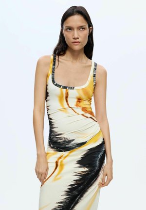 Femme aux longs cheveux foncés portant une robe sans manches ajustée avec des motifs abstraits jaunes, noirs et blancs sur un fond blanc uni.