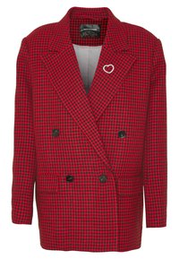 Veste croisée à motif pied-de-poule rouge et noir avec des boutons noirs et une petite épingle en forme de cœur argenté sur le revers gauche.