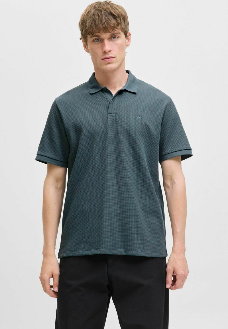 Koszulka polo w ciemnym tealowym kolorze, wykonana z teksturowanej tkaniny, z kołnierzykiem i trzyguzikową plisą, krótkimi, podwiniętymi rękawami oraz małym logo.
