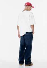 Ung person iført en rød hat, oversized hvid T-shirt, løse mørke jeans og brune sko, stående med hænderne i lommerne og vendt på siden.