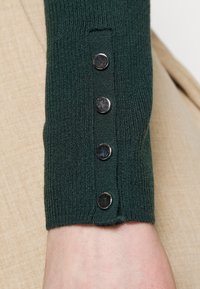 Zielona żeberkowa rękawica z czterema małymi okrągłymi srebrnymi zatrzaskami na nadgarstku na beżowym tle.