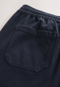 Tamsiai mėlynos sportinės kelnės pagamintos iš tekstūruotos medžiagos, turi elastingą juosmenį ir vieną atloštą kišenę su apvalia, kampuota forma.