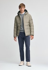 Giacca a vento beige con cappuccio, texture trapuntata, maglione grigio sotto, pantaloni blu navy e sneakers chiare.