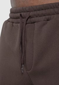 Nahaufnahme einer Person, die dunkelbraune Jogginghosen mit einem elastischen Bund und einer sichtbaren Zugkordel trägt, die vorne gebunden ist.