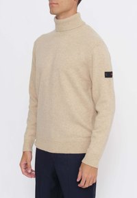 Maglione a collo alto beige realizzato in tessuto a maglia, con polsini e orlo a coste, con un dettaglio patch nero sulla manica.