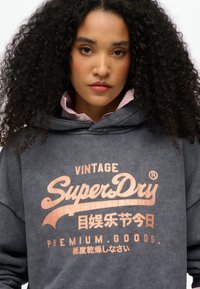 Sudadera gris oscura hecha de tela suave con un gran logo metálico de color cobre impreso que dice "Vintage SuperDry". Cuenta con capucha ajustable y un corte relajado.