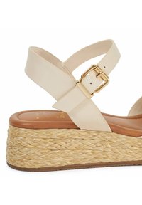 Beige leather sandal with crossed straps, gold buckle detail, and a woven jute platform sole. The footbed is tan with a smooth texture.