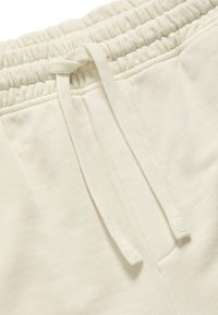 Primo piano della vita di pantaloni della tuta color crema con fascia elastica e cordini piatti annodati.