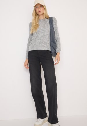 Pull en tricot gris, jeans noirs à jambes larges et baskets blanches. La mannequin porte un sac gris et un casquette claire. Tenue simple et décontractée.