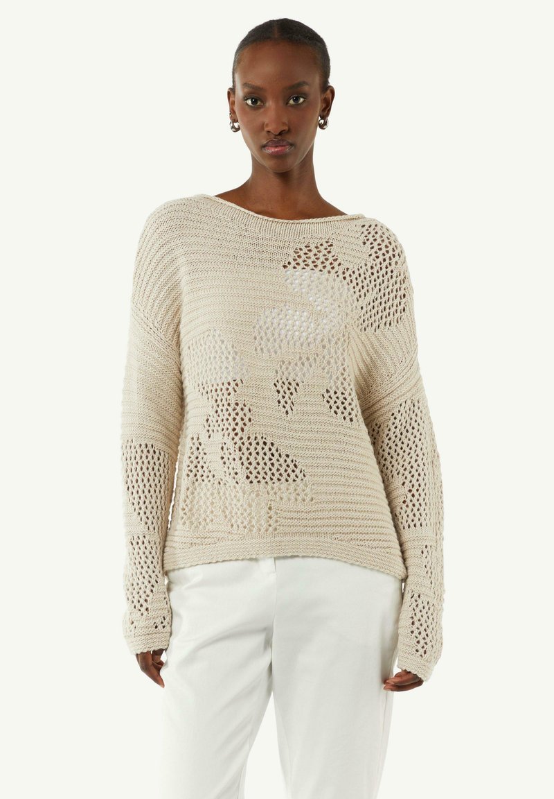 Strickpullover in hellbeige mit lockerer Passform, offener Webstruktur und floralen Ausschnitten, kombiniert mit weißen Hosen.