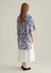 Kimono lungo colorato con motivo floreale, a maniche corte, realizzato in tessuto leggero. Indossato sopra una gonna bianca, abbinato a ballerine blu scuro.