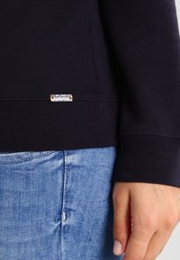 Marinblå sweatshirt med ribbade muddar, prydd med en silverlogotagg med orange detaljer. Tillsammans med ljusblå denimjeans.