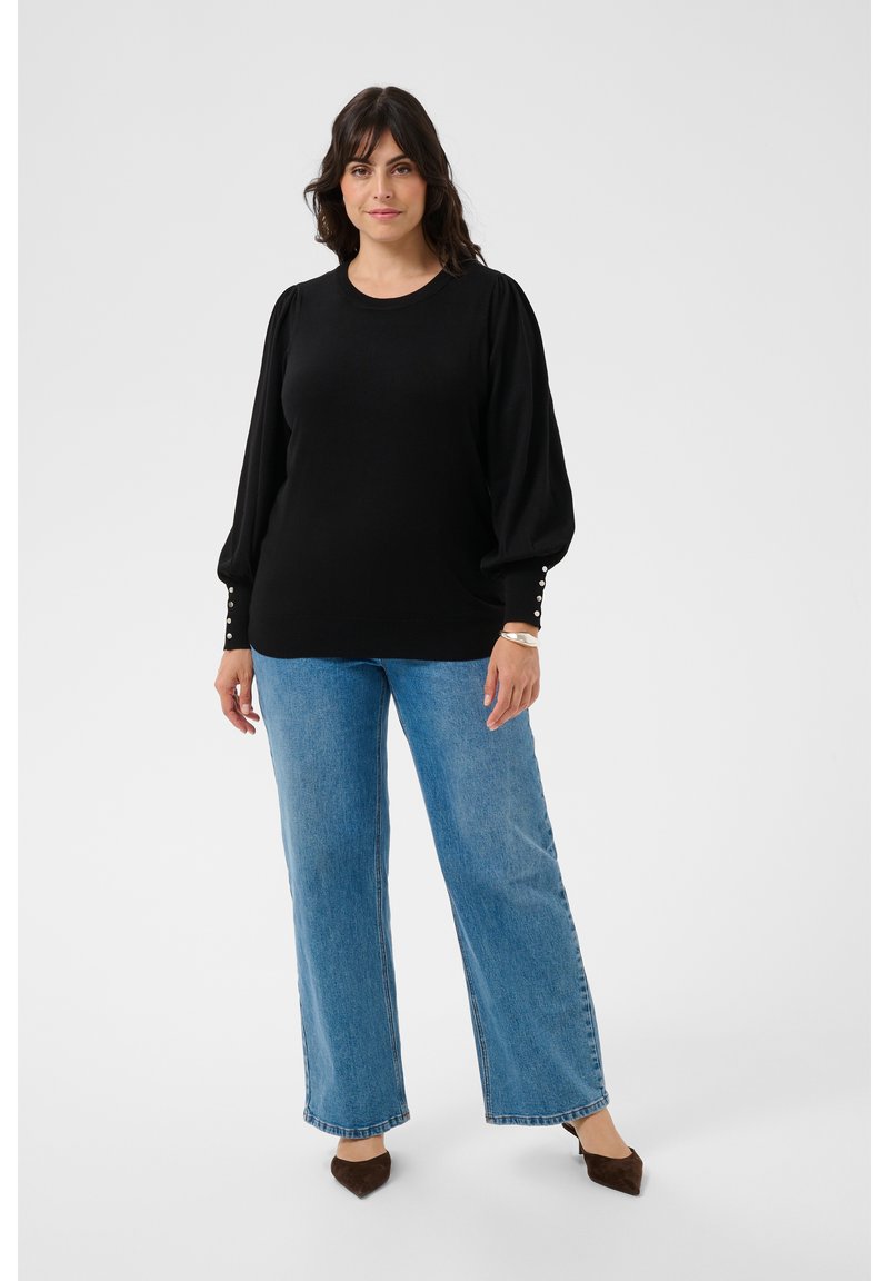 Femme en blouse noire à manches longues avec boutons en perle aux poignets, jean large bleu clair et chaussures marron pointues, debout sur un fond uni.