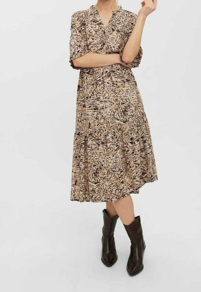 Femme portant une robe midi beige à motifs noirs, à manches courtes, et des bottines noires à bout pointu, se tenant les bras croisés.