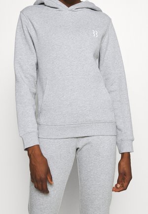 Osoba w jasnoszarej bluzie z kapturem i dopasowanych spodniach dresowych z małym, białym logo na lewej piersi, stojąca na jednolitym tle.