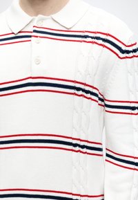 Pull polo blanc en tricot avec des rayures horizontales rouges et bleu marine et des détails en tricot torsadé sur les manches et les côtés.
