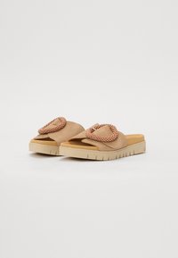 Sandali scivolati in suede beige con un cerchio decorativo di perline marroni sulla parte superiore. Suola in gomma spessa con una finitura testurizzata beige.