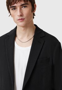 Schwarzer taillierter Blazer mit Reverskragen, strukturiertem Stoff, dezenten Taschendetails, getragen über einem weißen gerippten Rundhalshemd. Silberne Kettenhalskette.