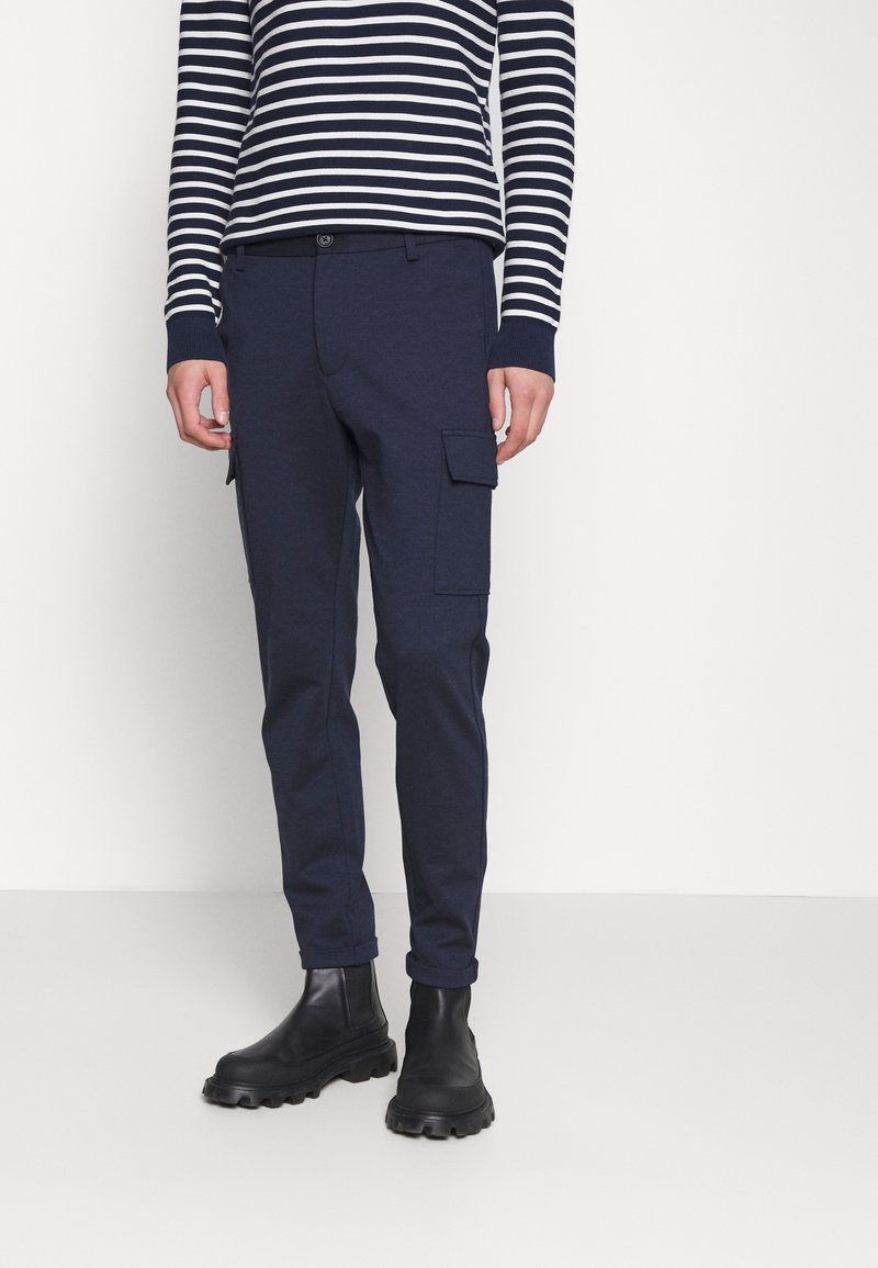Personne portant un pantalon cargo bleu marine, des bottes noires épaisses, et un haut à manches longues rayé bleu marine et blanc, debout sur un fond blanc uni.