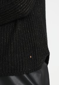 Svart stickad tröja med ribbat mönster; metalltråd i detaljerna och en liten knapp vid fållen. Den nedre kanten har en avslappnad passform.