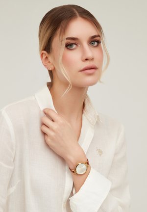 Young woman with blonde hair styled in loose strands, wearing a white button-up shirt and a gold watch with a brown leather strap.