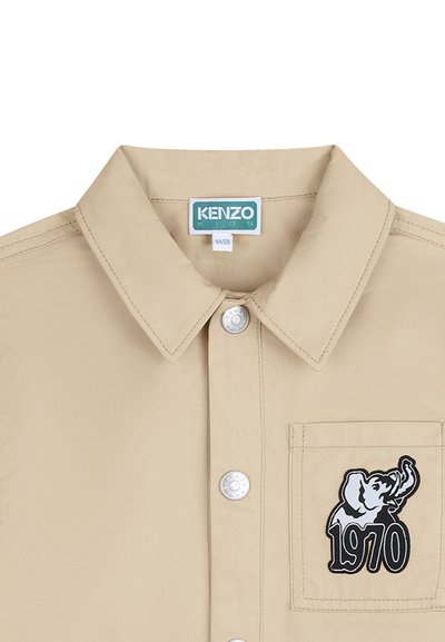 Beige overhemd met knoopsluiting en kraag, met een borstzak aan de linkerkant waarop een olifant is geborduurd en het jaartal "1970".