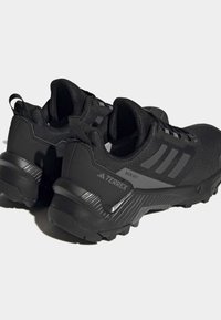 Baskets de sport noires avec une tige en mesh texturé et une semelle en caoutchouc, arborant trois rayures grises et le logo "TERREX RAIN.RDY" sur le côté.